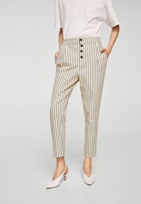 Pantaloni a righe beige con vita alta, dotati di chiusura a bottone, tasche laterali e gambe affusolate, abbinati a scarpe con tacco alto bianche.