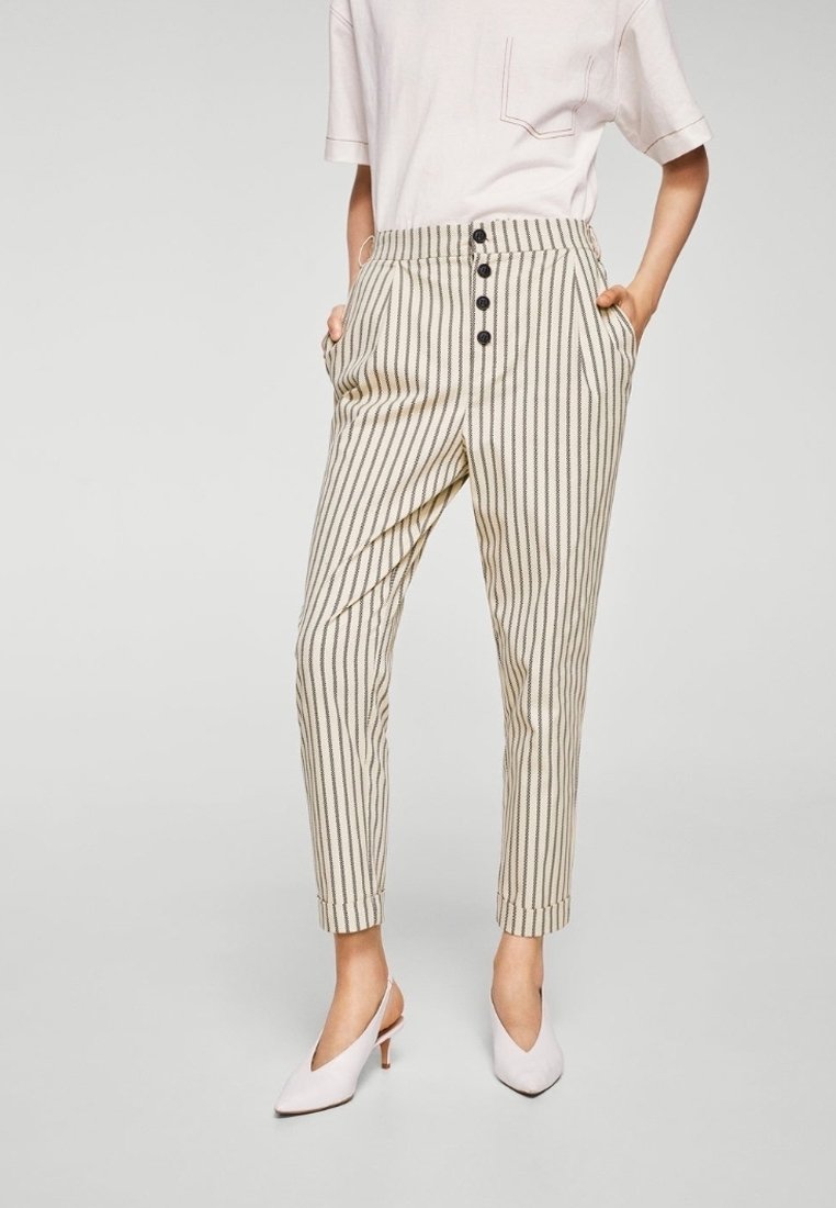 Pantaloni a righe beige con vita alta, dotati di chiusura a bottone, tasche laterali e gambe affusolate, abbinati a scarpe con tacco alto bianche.