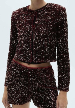 Mujer con chaqueta de manga larga con lentejuelas de color rojo oscuro y shorts de talle alto a juego frente a un fondo claro y liso.
