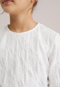 Haut en coton blanc présentant un motif brodé en relief avec un col rond et un tissu léger et doux. Finitions subtiles sur l'ensemble.