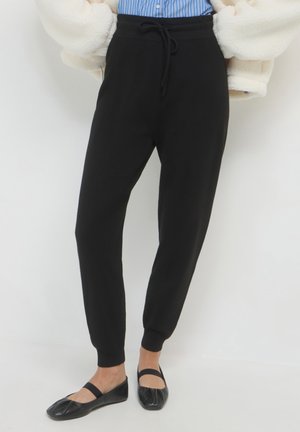 Jogging noir en coton avec une taille à cordon et des poignets de chevilles, associés à des chaussures noires plates. Le fond est entièrement blanc.