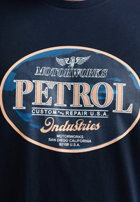 T-shirt en coton bleu marine avec un grand graphique circulaire affichant "PETROL" en lettres blanches et orange en gras, accompagné de texte supplémentaire et d'un emblème d'aigle.