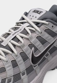 Közelkép egy szürke Nike tornacipőről fehér fűzőkkel, fekete pipa logóval, hálós felsőrésszel és "RONNAN SERIES" címkével a nyelvén.