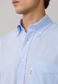 Camisa de botonada de color azul claro en tela de algodón. Cuenta con un cuello, botones blancos y un bolsillo en el pecho con una etiqueta. Textura suave.