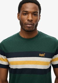 Man draagt een SuperDry T-shirt met groene, marineblauwe, grijze en gele strepen, kijkt recht vooruit met een neutrale uitdrukking tegen een witte achtergrond.