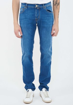 Uomo con jeans slim fit blu e sneakers bianche, in piedi con le braccia rilassate, tatuaggio visibile sull’avambraccio destro.