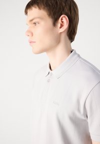 Polo shirt gris clair avec col côtelé, patte de boutonnage à deux boutons, et logo en relief subtil, doté d'une texture lisse et d'une coupe ajustée.