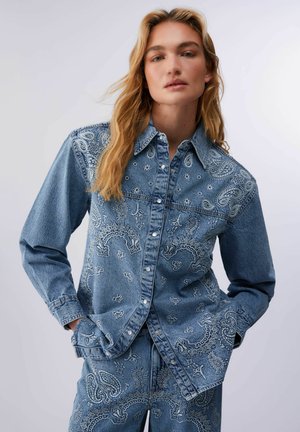 Jeune femme aux longs cheveux blonds portant une veste en denim bleue et un pantalon avec une broderie blanche à motifs cachemire, mains dans les poches, fond neutre.
