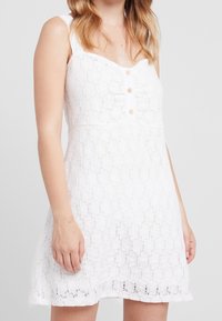 Vit spetsklänning med en kåpa som passar bra, knappfront med träknappar och en utsvängd kjol som visar blommönster och en texturerad yta.