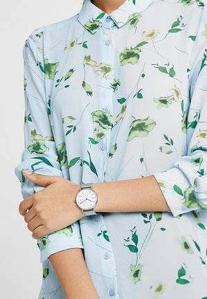 Mujer que lleva una camisa azul clara con estampado floral, abotonada y con las mangas remangadas, y un reloj con correa de malla plateada en la muñeca izquierda, brazos cruzados.