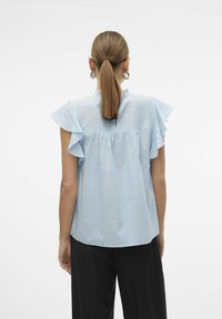 Blusa azzurra chiara con maniche a volant, texture a pois, collo alto e design con schiena arricciata. Abbinata a pantaloni neri.