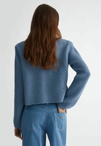 Sininen neulottu lyhyt neuletakki, jossa on teksturoitu kuvio ja pitkät hihat, yhdistetty vaaleansinisiin farkkuihin, joissa on takataskut ja ompeleen yksityiskohtia.