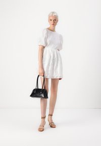 Λευκό φόρεμα με πούφ μανίκια και υφή λουλουδιών, με σούρα στη μέση. Το μοντέλο κρατά μια μικρή μαύρη δερμάτινη τσάντα με δύο χερούλια. Μπεζ σανδάλια με ζώνες.