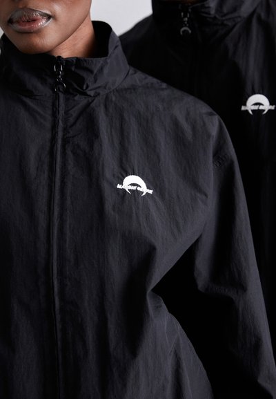 Veste légère noire avec un col haut. Présente un logo blanc sur la poitrine gauche. Texture lisse, fermeture éclair et design minimaliste.