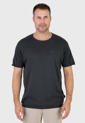 Mann mit kurzen braunen Haaren, der ein schwarzes kurzärmeliges T-Shirt und beige Hosen trägt, steht vor einem schlichten, hellen Hintergrund.