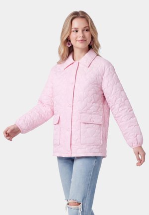 Femme portant une veste matelassée rose clair avec des motifs en forme de cœur et de grandes poches avant, associée à un jean bleu clair déchiré.