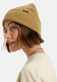 Beige ribbed knit beanie with a pointed top and a black "ROXY" logo patch. The model has short hair and wears a light-colored top.