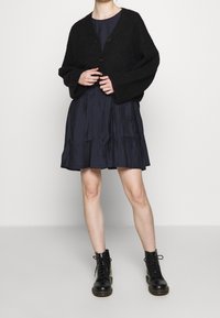 Eine Frau trägt eine schwarze Knopfkardigan, ein knielanges marineblaues Kleid und schwarze Schnür-Ankle Boots und steht vor einem schlichten weißen Hintergrund.