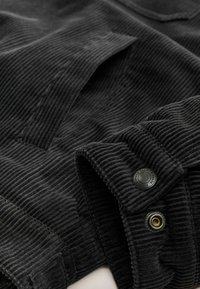 Tejido de pana negro con textura acanalada, que cuenta con bolsillos y acentos de botones metálicos. El detalle en primer plano muestra la costura de las costuras y el cierre con botón.