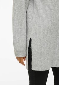 Roka, kas atpūšas blakus gaiši pelēkam adītam džemperim ar sānu šķelumu, uzvilkts virs melnām biksēm.