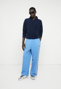 Dunkelblauer Strickpullover mit Kragen, kombiniert mit hellblauen Sweatpants mit schwarzem Seitenstreifen und Logo. Weiße und blaue Sneakers runden den Look ab.