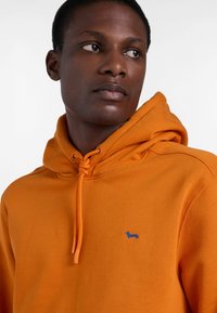 Pomarańczowa bluza z kapturem, wykonana z miękkiego materiału, z kapturem ze sznurkiem, małym niebieskim haftowanym logo i luźnym krojem.