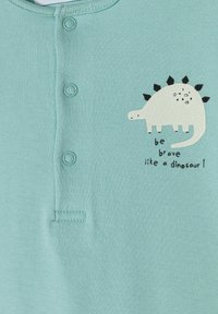 Chemise à manches courtes vert clair ornée d'un graphique de dinosaure blanc avec des épines noires et du texte "sois brave comme un dinosaure" en dessous.