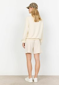 Beige souprava mikiny a šortek s uvolněným střihem; strukturovaná látka. Bílé tenisky a kšiltovka s leopardím vzorem doplňují outfit.