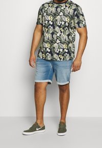 Camiseta de estampado floral en azul marino oscuro con hojas verdes y flores blancas, combinada con pantalones cortos de denim azul claro y zapatillas verde oliva.