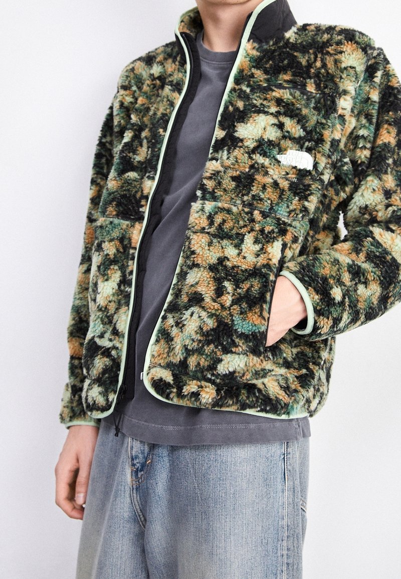 Flauschige Jacke mit grün-braunem Muster, mit Reißverschluss vorne, kontrastierenden hellgrünen Akzenten und Fronttaschen. Wird über einem grauen T-Shirt getragen.