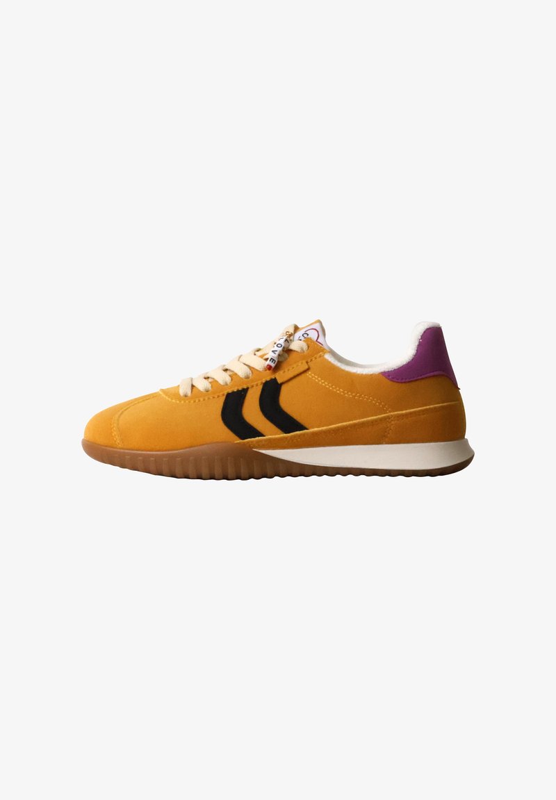 Baskets en daim jaune avec des rayures en chevron noires, des lacets crème, un insert violet au talon et une semelle en caoutchouc marron, vues de côté sur fond blanc.