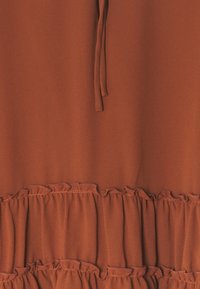 Rostfärgad tyg med en slät textur, med volangdetaljer i botten och en snörning vid halsringningen.