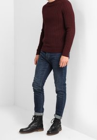 Uomo che indossa un maglione bordeaux a treccia, jeans blu scuro arrotolati e stivali di pelle neri con lacci, in piedi contro un muro bianco.