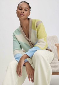 Neulejakku, jossa on vihreät, keltaiset, beiget ja siniset raidat, syvällä V-kaula-aukolla ja ribbaetuilla hihoilla, yhdistettynä kermaisiin housuihin.