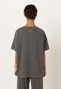 Ženska z kodrastimi lasmi spetimi v figo, oblečena v ohlapno temno sivo kratko srajco in hlače, stoji obrnljena proti enostavni svetli podlagi.