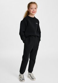 Zwarte sweatshirt en joggers van zacht materiaal, met een logo op de borst en een trekkoord in de taille. Gecombineerd met zilveren sneakers.