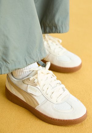 Person iført beige Puma-sneakers med hvide snørebånd, hvide sokker og grønne bukser, der står på en gul overflade.