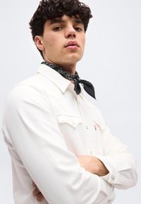 Νεαρός άνδρας με σγουρά μαλλιά, που φοράει λευκό πουκάμισο και μαύρο μαντήλι με σχέδια στο λαιμό, με σταυρωμένα τα χέρια, κοιτάζοντας ελαφρώς προς τα πάνω.