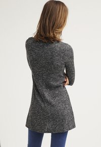 Cardigan lungo grigio scuro con un motivo a maglia strutturato, dotato di un orlo curvo e maniche lunghe. Indossato sopra jeans blu, visto da dietro.