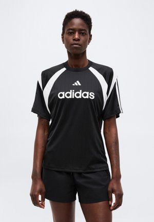 Person med kort hår iført sort Adidas sportstrøje med hvide striber og logo, kombineret med sorte shorts, stående mod en ensfarvet baggrund.