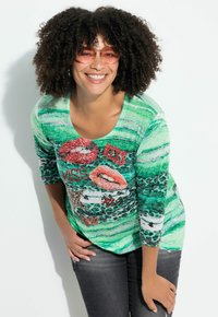 Zielony, w paski top z długim rękawem z grafiką ust i akcentami tekstowymi. Posiada okrągły dekolt oraz czarne, białe i panterkowe wzory.