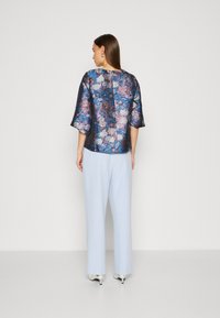 Haut à motifs floraux bleu et violet avec des manches à trois-quarts, associé à un pantalon bleu clair et des talons blancs, vu de derrière.