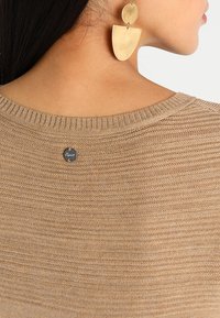 Pull en tricot beige avec un col côtelé, arborant une petite étiquette logo circulaire à l'arrière. Grandes boucles d'oreilles pendantes en métal doré visibles.