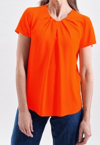 Blusa arancione brillante a maniche corte con scollo plissettato e tessuto liscio. Abbinata a jeans in denim scuro, che mostrano una vestibilità rilassata.