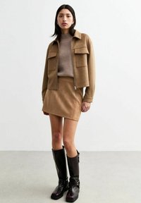 Braune Wildleder-Cropped-Jacke mit zwei Fronttaschen, kombiniert mit einem passenden Minirock und knielangen schwarzen Stiefeln. Darunter ein strukturierter Strickpullover.