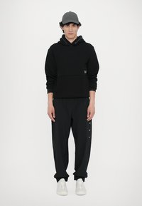 Felpa nera con cappuccio e tasca frontale, abbinata a pantaloni jogger neri con stampa del logo bianco. Il modello indossa un cappellino grigio e sneakers bianche.
