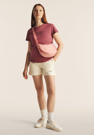 T-shirt rosa con stampa "Freddy MOV", pantaloni corti color crema e borsa a tracolla rosa. Sneakers bianche e calzini alla caviglia. Design semplice e casual.