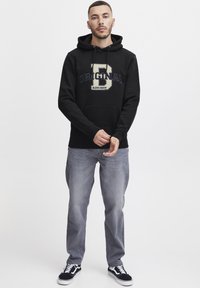 Black hoodie with "ORIGINAL" and logo print in cream and navy, kangaroo pocket, paired with light blue jeans and black sneakers.