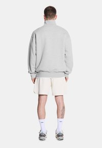 Młody mężczyzna widziany z tyłu, ubrany w szary bluzę, kawałki kremowych szortów i trampki, z widocznym tatuażem na nodze.