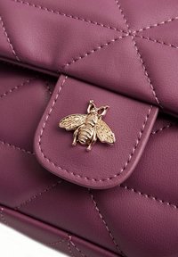 Borsa in pelle viola trapuntata con chiusura a farfalla dorata. Presenta una texture liscia e cuciture tono su tono, evidenziando un design moderno.